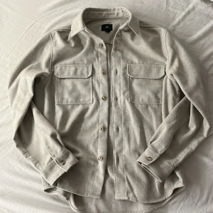 Beige overshirt från H&M i bomull - Säljer en stilren beige overshirt från H&M i mjuk bomull. Skjortan har klassisk krage, två stora bröstfickor med lock och knäppning framtill. Perfekt lager-på-lager-plagg med lång ärm och snygg passform.