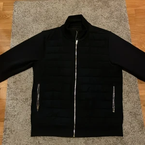 Mörkblå quiltad bomberjacka Next - Snygg mörkblå bomberjacka från Next med quiltad framsida och silverfärgade dragkedjor. Jackan har ribbad krage och raka ärmar, perfekt för dig som gillar stilren och modern look. Passar dig som vill ha en jacka med lite edge och coola detaljer.