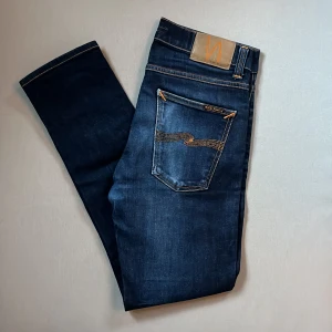 Mörkblå Nudie Jeans Lean Dean W30 L30 - Snygga mörkblå jeans från Nudie Jeans, modell Lean Dean. Klassisk femficksdesign med orange kontrastsömmar och ikonisk broderad bakficka. Tillverkade i 99% bomull och 1% elastan för en bekväm passform. Perfekta för dig som gillar stilrena jeans med modern känsla.