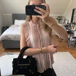 Ljusrosa blus med volang från Zara - Supersöt ljusrosa blus från Zara med volangdetaljer längs axlar och krage. Blusen är ärmlös och har plisserade partier framtill samt knappar. Materialet är lätt och luftigt, perfekt för vår och sommar. Snygg att styla med jeans eller kjol! Toppen är i nyskick och endast använd en gång.