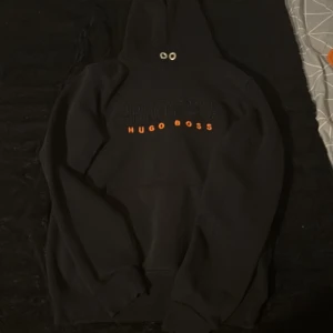 Svart hoodie från Hugo Boss - Svart hoodie från Hugo Boss med broderad logga i svart och orange text på bröstet. Klassisk huva med snörning och magficka framtill. Perfekt för dig som gillar stilrena och snygga streetwear-plagg.