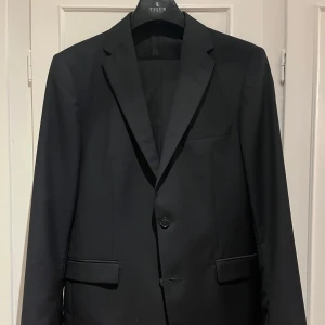 Svart kostym - Tiger of Sweden - Klassisk svart kostym från Tiger of Sweden i modell HENRIE. Kavajen har två knappar, smala slag och fickor med lock. Byxorna är raka och stilrena. Kostymen är tillverkad i 100% ull och har ett elegant, tidlöst snitt som passar perfekt till mer uppklädda tillfällen. Se bild på den lilla tygskada som är  på vänster ärm! (Köparen står för frakten).