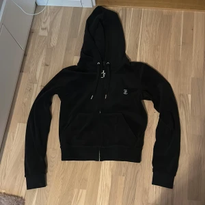 Svart Juicy Couture tjocktröja  - Svart hoodie från Juicy Couture i mjuk sammet med glittrande Juicy-text på ryggen. Dragkedja framtill med snygg JC-berlock, huva med snörning och känguruficka. Perfekt för dig som vill ha en ikonisk och trendig look.