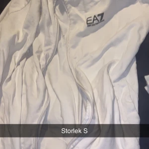 Vit tröja från EA7 Emporio Armani - Säljer en stilren vit tröja från EA7 Emporio Armani med dragkedja framtill och diskret logga på bröstet. Tröjan har en clean look och passar perfekt för dig som gillar sportig och modern stil.