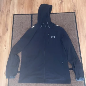 Svart vindjacka från Under Armour - Svart vindjacka från Under Armour med huva och hel dragkedja. Jackan har reflekterande detaljer på axlarna, bröstficka med dragkedja och logga på bröstet. Perfekt för blåsiga dagar och träning utomhus.