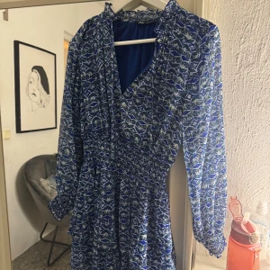 Blå mönstrad omlottklänning från Zara - Supersnygg blå omlottklänning från Zara med vitt och grönt mönster. Klänningen har lång ärm med resår vid ärmslut, markerad midja och v-ringning. Lätt och luftig känsla, perfekt för varmare dagar. Säljer då den inte kommer till användning längre.
