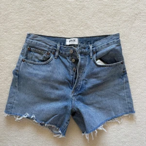 Blå jeansshorts från Agolde - Säljer ett par klassiska blå jeansshorts från Agolde med hög midja och råa, fransiga benslut. Shortsen har fem fickor, knappgylf och bälteshällor. Perfekta för sommaren och ger en avslappnad vibe.