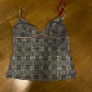 Rutig linnetopp med rosa detaljer - Supersnygg linnetopp från Misstriumph med rutigt mönster i svart, vitt och grått. Toppen har tunna, justerbara axelband i ljusrosa och en markerad söm under bysten. Perfekt för dig som gillar trendiga och stilrena plagg med en twist.