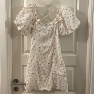 Vit blommig kortklänning H&M XXS - Supersöt vit kortklänning från H&M med små rosa blommor, puffärmar och fina rosettdetaljer vid axlarna. Klänningen har korsade band i ryggen och är gjord i ett lätt bomullstyg – perfekt för sommarens alla tillfällen!