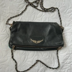 Svart axelväska från Zadig & Voltaire - Cool svart axelväska i skinn från Zadig & Voltaire med kedjerem och snyggt vingemblem i metall framtill.                                                                   Köpt 2018. Använd i ca 2 år. Ganska sliten på kanten och i lädret men går att lämna in till skomakare och putsa lädret. Silverdetaljerna har tappat färg. I helheten fin men när man tittar nära ser man defekter! Kvitto finns inte kvar men den är köpt på Zadig bu