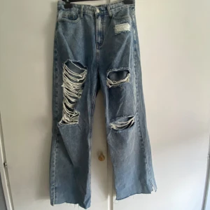 Blå slitna wide jeans med hål - Ett par ljusblå jeans med rejält slitna detaljer och stora hål på båda benen. Modellen är wide fit med hög midja och klassisk femficksdesign. De är perfekt för en avslappnad men stilsäker look. De är i nyskick - endast använda en gång - och har inga fläckar, hål (förutom de som ska vara där) eller andra skador.