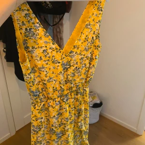 Gul blommig klänning Samsøe Samsøe - Superfin gul klänning från Samsøe Samsøe med blommigt mönster i blått och vitt. Klänningen är ärmlös, har v-ringning och markerad midja. Perfekt för sommarens alla tillfällen och ger en riktigt fräsch vibe.
