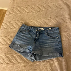 Blå jeansshorts från H&M, strl 36 - Säljer ett par klassiska blå jeansshorts från H&M i storlek 36. De har normal passform, fem fickor och uppvikta benslut. Perfekta för sommaren och lätta att matcha med allt. Stängs med dragkedja och knapp framtill.