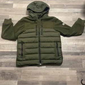 Olivgrön pufferjacka med huva - Olivgrön pufferjacka med stickade ärmar och huva. Jackan har flera dragkedjeförsedda fickor framtill och quiltad kropp. Materialmix med vadderad polyester och stickade partier. Perfekt för kyliga dagar och har en sportig vibe.enda effekten på jackan är på sista bilden, hör av er vid frågor!