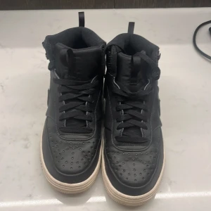 Nike Air Force 1 Mid svart/vit - Svarta Nike Air Force 1 Mid sneakers med vit sula. Klassisk design med perforerad tå, snörning och Nike-logga på sidan. Ovandel i skinn och syntet, vadderad plös och häl för extra komfort. Perfekta för dig som gillar streetstyle och tidlös look.