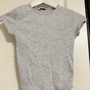 Grå ribbad t-shirt från Bershka - En basic grå ribbad t-shirt från Bershka i storlek XS. T-shirten har korta ärmar, rund halsringning och är tillverkad i mjuk bomullsmix. Perfekt att styla med jeans eller kjol för en clean och enkel look.