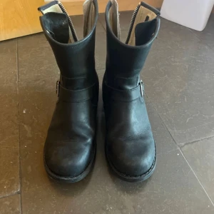 Svarta Johnny Bulls boots i skinn - Säljer ett par svarta boots från Johnny Bulls i äkta skinn. De har grov sula, rund tå, spännen i silver och dragkedja baktill. Klassisk modell med robust känsla, perfekta till jeans eller kjol för en cool look. Ena dragkedjan är sönder men går att fixa genom att köpa en ny eller om man man tar i hårt. 