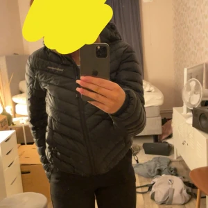 Svart pufferjacka från Peak Performance - Säljer en svart pufferjacka från Peak Performance med huva och dragkedja framtill. Jackan har en stilren look med lätt vaddering och diskret logga på bröstet. Perfekt för kyliga dagar och enkel att matcha med olika outfits. Några små hål där bak men inget märkvärdigt 