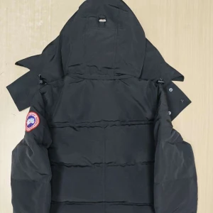 Svart dunjacka från Canada Goose - Säljer en svart dunjacka från Canada Goose med stor huva och klassisk logga på ärmen. Jackan har knäppning framtill, diskreta sidofickor och en quiltad design som håller värmen. Perfekt för kalla vinterdagar och riktigt snygg streetstil.