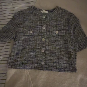 Grå tweed kofta med knappar - Säljer en kort grå kofta i tweed-look med rund halsringning och stora dekorativa knappar framtill. Koftan har två fickor med knappar på bröstet och korta ärmar. Perfekt för dig som gillar klassisk och trendig stil.