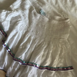 Vit volangkjol med paljettdetaljer - Supersöt vit kortkjol med volanger och glittriga paljettdetaljer i grönt, lila och rosa längs kanterna. Kjolen har smock i midjan för en bekväm passform och är perfekt för dig som gillar en lekfull och trendig stil.