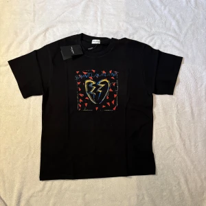 Svart Saint Laurent t-shirt med tryck - Riktigt fin Svart t-shirt från Saint Laurent med färgstarkt tryck på bröstet som visar ett blixtformat hjärta och röda trianglar. T-shirten har klassisk passform, rund hals och är tillverkad i mjuk bomull. Perfekt statement-plagg för dig som gillar streetstyle. Sitter som M men ni får bedöma själva, modellen är 179 65kg och brukar bära S