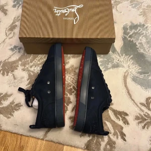 Christian Louboutin svarta sneakers - Snygga svarta sneakers från Christian Louboutin i mocka med klassisk röd sula och broderad logga på hälen. Skorna har snörning och rund tå, vilket ger en clean och exklusiv look. Perfekta för dig som vill sticka ut med lyxiga detaljer.