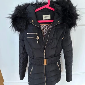 Svart pufferjacka med fuskpäls från Roco Baroco - Svart pufferjacka från Roco Baroco med fuskpäls på huvan, gulddetaljer och flera dragkedjor. Jackan har bälte i midjan och quiltad design. Perfekt för kalla dagar och ger en snygg siluett.