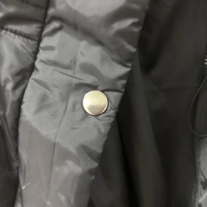 Svart  puffväst med huva - Säljer en svart,  puffväst med stor huva och dragsko i midjan. Jackan har blanka silverfärgade knappar och dragkedja framtill. Perfekt för lager-på-lager och chill vilket ger en modern vibe. Storlek XL  (katt finns i hemmet) 