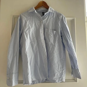 Ljusblå skjorta från H&M - En klassisk ljusblå skjorta från H&M med lång ärm, bröstficka och knappar framtill. Skjortan har en rak passform och är tillverkad i mjuk bomull, perfekt för en clean och enkel stil.