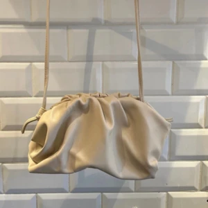 Beige axelväska med snörning - Trendig beige axelväska i mjukt skinnliknande material med cool snörning upptill. Väskan har en avslappnad, puffig form och en smal axelrem som gör den enkel att bära över axeln. Perfekt för dig som gillar stilrena accessoarer med en twist.