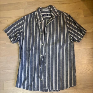 Randig kortärmad skjorta strl 164 - Snygg blå och vit randig linneskjorta från Jack & Jones. Skjortan har korta ärmar, klassisk krage och knäppning framtill. Perfekt för en chill och stilren look. 