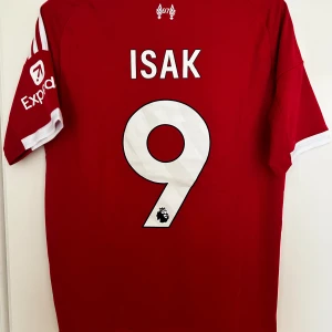 Liverpool FC röd matchtröja Isak #9 - Liverpool FC fotbollströja i röd färg med vita detaljer och tryck. Adidas-logga på bröstet, Standard Chartered som sponsor och Expedia på ärmen. Spelarnamn ISAK och nummer 9 på ryggen. Tillverkad i lätt, ventilerande material med klassiska vita ränder på axlarna.