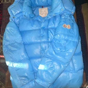 Blå Moncler pufferjacka med huva size L passar m - Säljer en blå pufferjacka från Moncler med avtagbar huva och klassisk logga på ärmen. Jackan är vadderad och har en glansig finish, perfekt för kalla dagar. Flera fickor och hög krage för extra värme. Riktigt snygg och trendig modell!