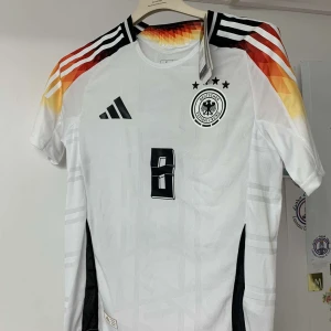 Tyskland Kroos  fotbollströja Adidas - Snygg Tyskland fotbollströja från Adidas med Kroos och nummer 8 på ryggen. Vit bas med svarta detaljer, svarta och vita ränder på axlarna samt coola inslag av rött, orange och gult upptill. Broderat landslagsmärke och stjärnor på bröstet. Tillverkad i lätt och ventilerande material.