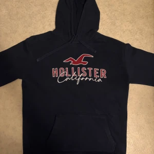 Mörkblå Hollister hoodie med tryck - Snygg mörkblå hoodie från Hollister med stort rött och vitt tryck på bröstet där det står 'Hollister California'. Tröjan har huva med snörning och en klassisk magficka. Perfekt för chill dagar och streetstyle.