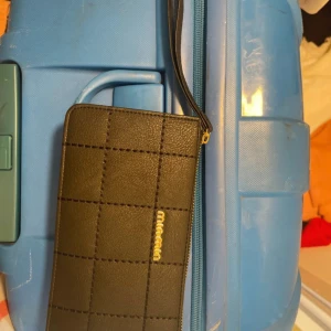 Svart plånbok från Missoni - Svart plånbok från Missoni med rutigt mönster och guldfärgad logga. Plånboken har flera fack, dragkedja och en praktisk handledsrem. Tillverkad i skinnliknande material och har stilren design som passar till det mesta.