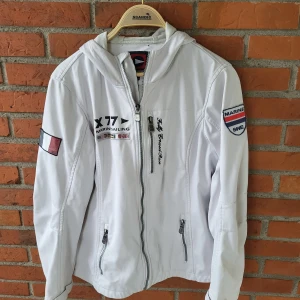 Vit vindjacka från Marine Sailing Storlek L  - Vit vindjacka från Marine Scandinavian Sportswear med broderade flaggor, tryck och patchar. Jackan har huva med dragsko, flera dragkedjeförsedda fickor och sportig look. Perfekt för dig som gillar segling eller vill ha en clean stil.