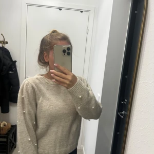 Beige stickad tröja med pärlor - Superfin beige stickad tröja med pärldetaljer på ärmarna. Tröjan har rund halsringning, ribbade muddar och en lite loose passform. Perfekt för dig som gillar en söt och stilren look med det lilla extra.