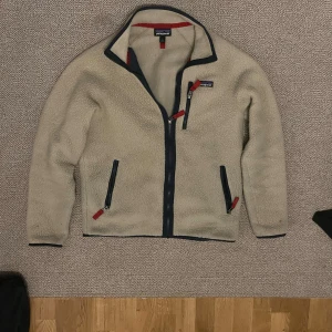 Beige fleecejacka från Patagonia - Mysig beige fleecejacka från Patagonia med marinblå detaljer och röda dragkedjor. Jackan har tre fickor med dragkedja och hög krage. Perfekt för kyliga dagar och enkel att matcha med det mesta. 