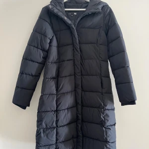 Svart lång pufferjacka från 157 - Lång svart pufferjacka från 157 med huva. Jackan har dragkedja och tryckknappar framtill, samt långa ärmar. Perfekt för kalla dagar och har en clean, stilren look❣️