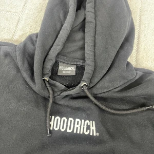 Svart hoodie från Hoodrich med logga - Svart hoodie från Hoodrich med vit logga på bröstet. Klassisk känguruficka framtill, justerbar huva med snörning och mjukt material. Perfekt för dig som gillar streetwear och vill ha en stilren look.