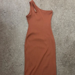Rostfärgad one shoulder klänning H&M - Säljer en snygg rostfärgad ribbad klänning från H&M. Modellen är tight och har en cool one shoulder-design med en extra cutout vid axeln. Klänningen är kort och gjord i ett stretchigt material som sitter skönt på kroppen. Använd endast en gång, så gott som ny, storlek S men funkar för M även, då den är stor i passformen. 