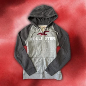 Grå zip hoodie med dragkedja från Hollister S/M - Grå zip hoodie med dragkedja från Hollister med vit text ’hollister’ och röd märkan | Size S/M | Längd: 66cm Armlängd: 68cm