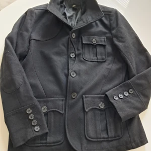 Svart ullkavaj från H&M, strl 52 - Stilren svart kavaj från H&M i ullmix med snygga fickdetaljer och knappar framtill. Klassisk krage, tre fickor med lock och dekorativa knappar vid ärmslut. Perfekt för dig som gillar en dressad look med modern touch.