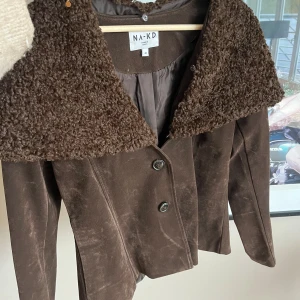 Brun teddyjacka från NA-KD, stl 40 - Snygg brun teddyjacka från NA-KD med stor, fluffig krage i teddy och knappar framtill. Jackan har en croppad passform och är tillverkad i mjuk mockaliknande polyester. Perfekt för dig som vill sticka ut med en unik och trendig look.