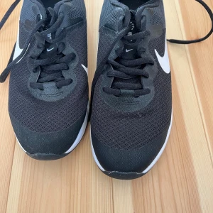 Svarta Nike sneakers  - Snygga svarta Nike sneakers med vit swoosh på sidorna. Ovandelen är i mesh för bra andningsförmåga och skorna har klassisk snörning. Perfekta för dig som gillar sportig stil och vill ha bekväma skor till vardagen.