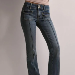 Blå bootcut jeans från Nelly - Snygga blå bootcut jeans från Nelly med dubbla knappar framtill. Jeansen har markerade sömmar längs benen och två stora fickor framtill. Tillverkade i klassiskt denimtyg med en cool passform som ger en trendig siluett. Jeansen har en liten slitning bredvid de två knapparna högst upp som man kan se på bilden.