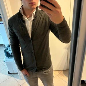 Grön merinoullskofta från WE XS - Snygg mörkgrön kofta från WE i 100% merinoull. Modellen har klassisk krage, knappstängning framtill och en bröstficka. Långärmad och tunn, perfekt för lager-på-lager. Passar dig som gillar stilrena och bekväma plagg. Bra skick.