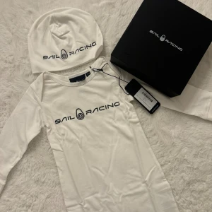 Sail Racing babyset helt nytt i presentbox 12–18 mån - Sail Racing babyset i vitt jumpsuit och mössa. Setet innehåller en långärmad mjuk jumpsuit med tryckknappar längs benen och med loggan, samt en matchande mössa till. Storlek 12–18 månader 100% Bomull Passar till både kille och tjej.  🌟 Helt ny med tag kvar! 🌟 Levereras i original presentförpackning, perfekt som present. Finns fler färger och olika storlekar i mina annonser 😍